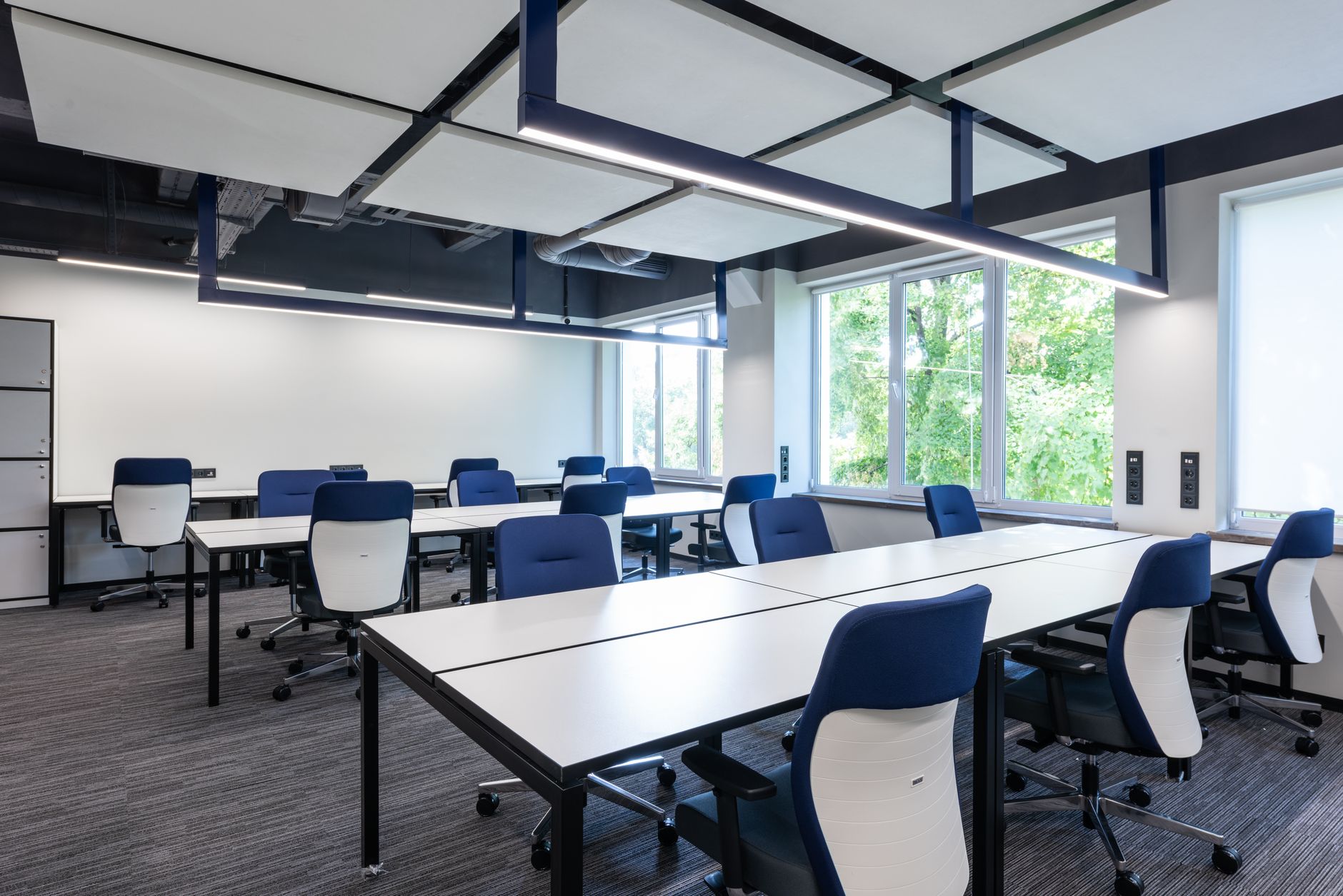 Open space vide avec tables blanches, fauteuils de bureau bleus et blancs, panneaux acoustiques au plafond et fenêtres ouvrant sur des arbres.