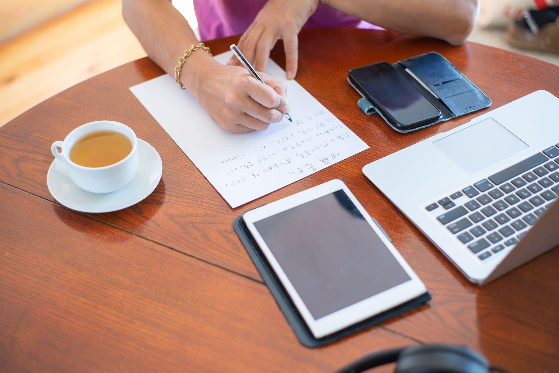 Personne écrivant sur une feuille, entourée d’un ordinateur portable, d’une tablette, d’un smartphone et d’une tasse de thé sur une table en bois.