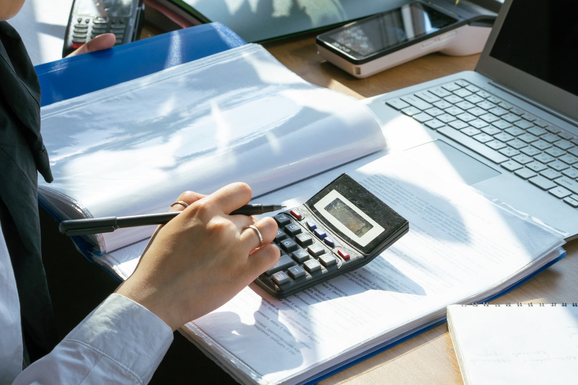 Une main utilise une calculatrice sur des documents, à côté d’un ordinateur portable, d’un classeur bleu et d’un carnet sur un bureau.