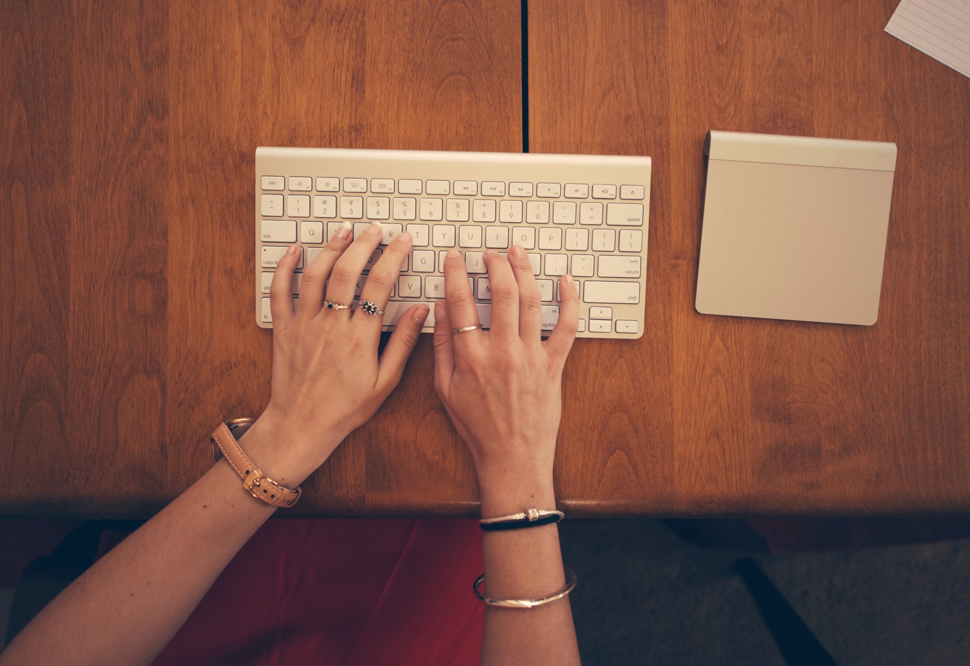 Vue de dessus de deux mains qui tapent sur un clavier sans fil blanc posé sur un bureau en bois, à côté d’un pavé tactile.